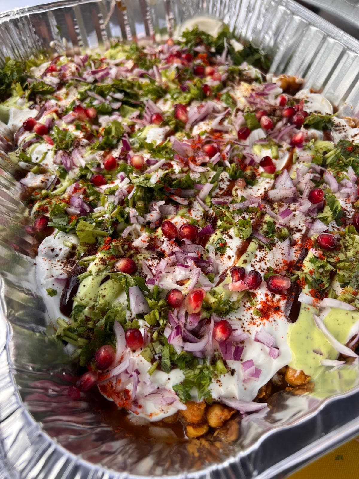 A large tray of chana chaat topped with yoghurt, tamarind and coriander chutneys, red onion, pomegranate seeds and chilli powder.