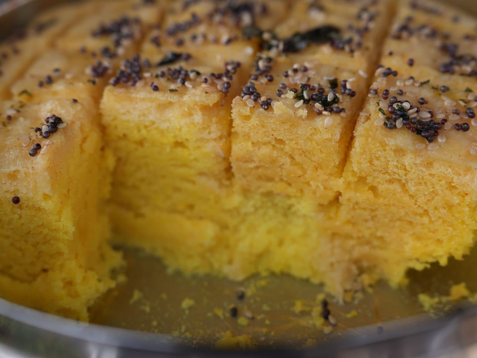 Dhokla cut into squares in the steamer tray, the first few pieces already lifted out, mustard seeds scattered across the top.