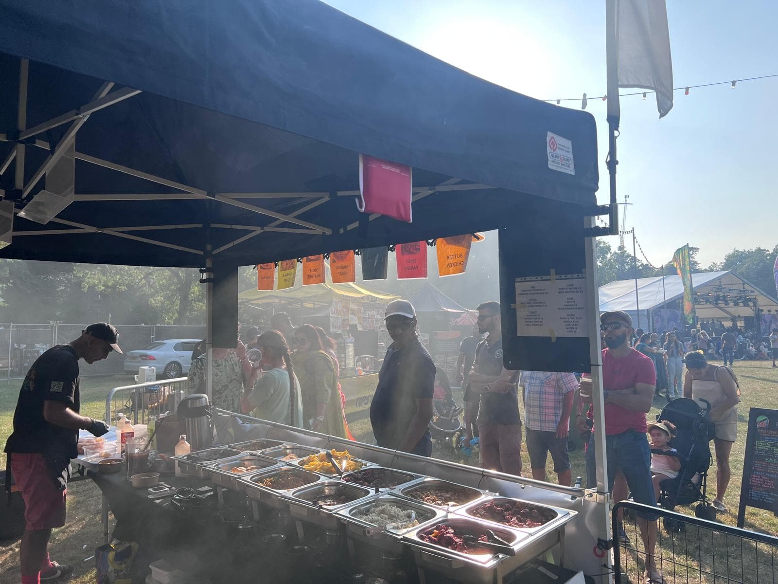 A cook plating up behind a row of steaming curry trays at the Karoga Corner stall, with festival-goers queuing along the counter in low afternoon sun.
