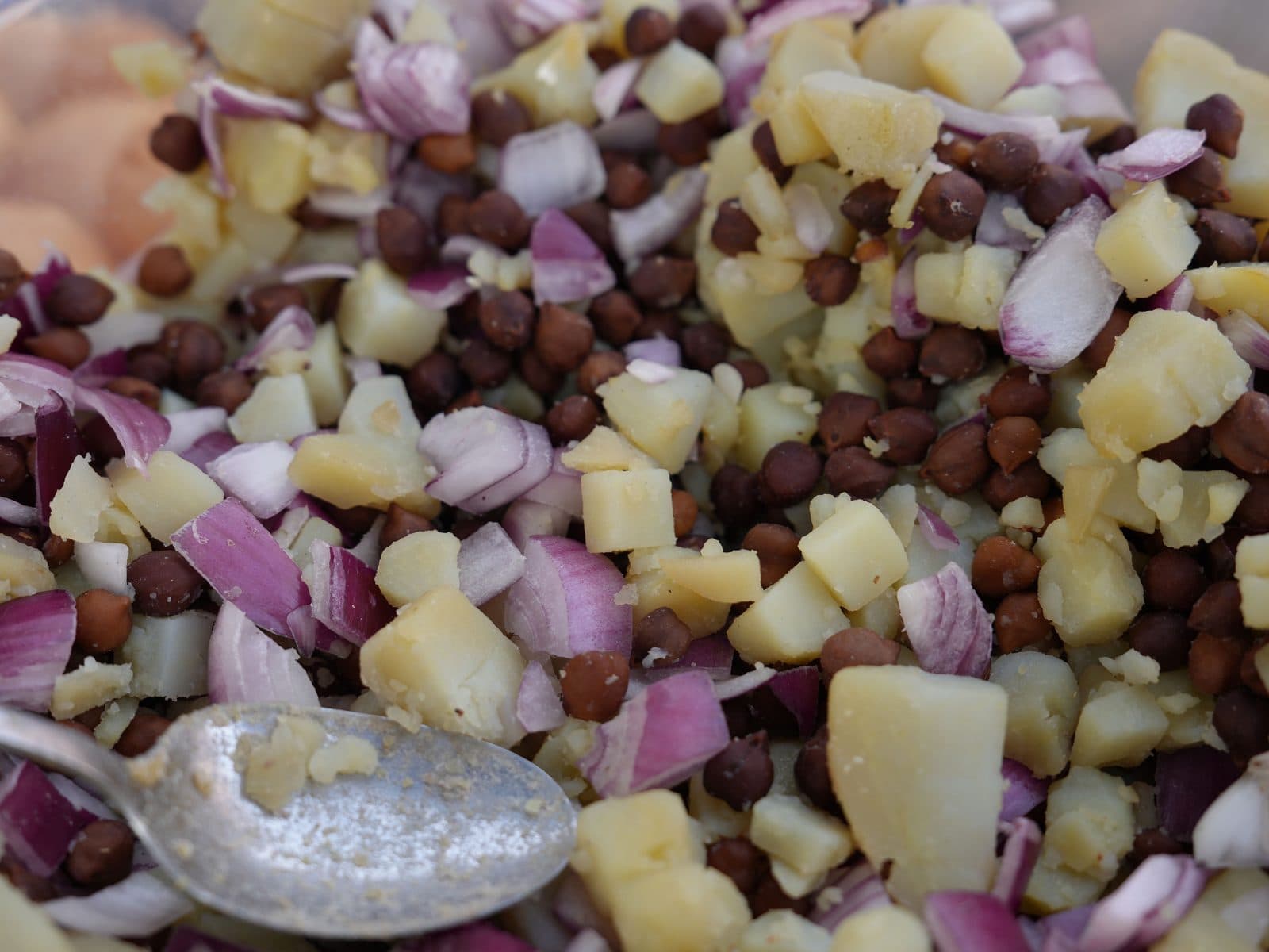 A close-up of kala chana and diced potato tossed with red onion, a serving spoon resting in the tray.