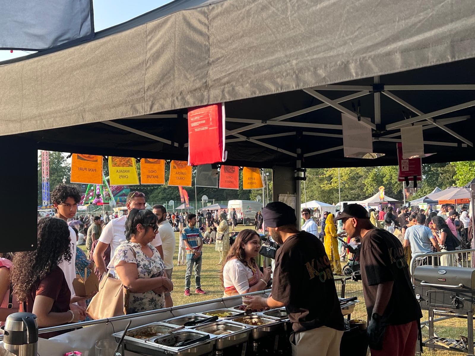 Two Karoga Corner cooks serving from the hot counter while a crowd fills the field behind, under a row of hanging menu flags for pani puri, masala chips and chole bhature.