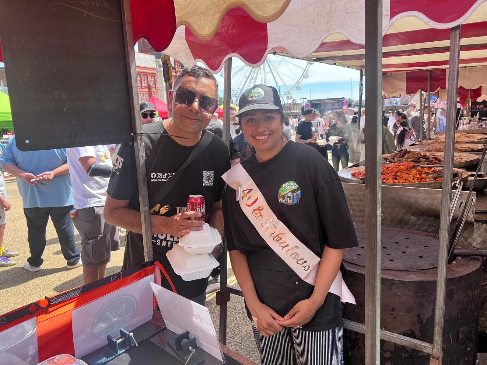 The musician Panjabi MC and a member of the Karoga Corner team, in a branded cap and a 40th-birthday sash, at the stall on a busy mela day, with trays of grilled meat on the tawas beside them and a big wheel above the crowd behind.