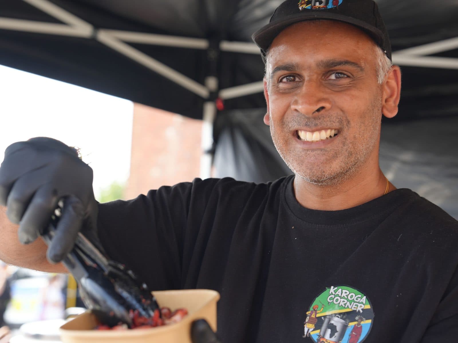 A member of the Karoga Corner team in a black cap and branded shirt, gloved and holding tongs, grinning at the camera while plating up under the gazebo.