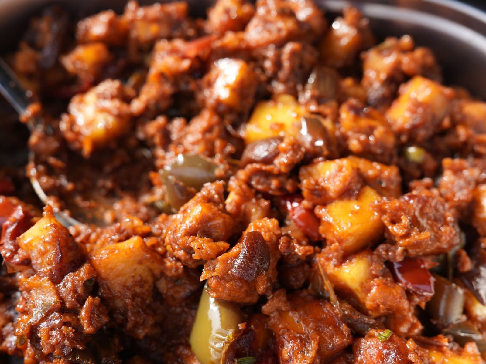 A close-up of potato and meat in a dark red masala with onion and green chilli, a serving spoon lifting through it.
