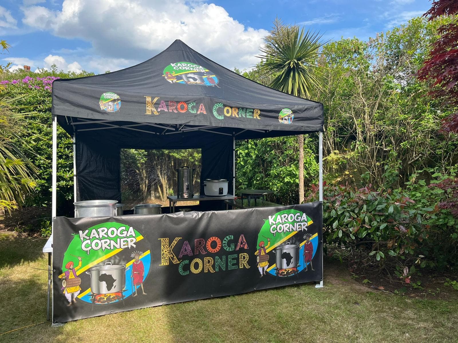 The Karoga Corner stall fully dressed under a clear sky, with hand-painted lettering across the canopy, the logo banner along the front and two karoga pots waiting on the counter.