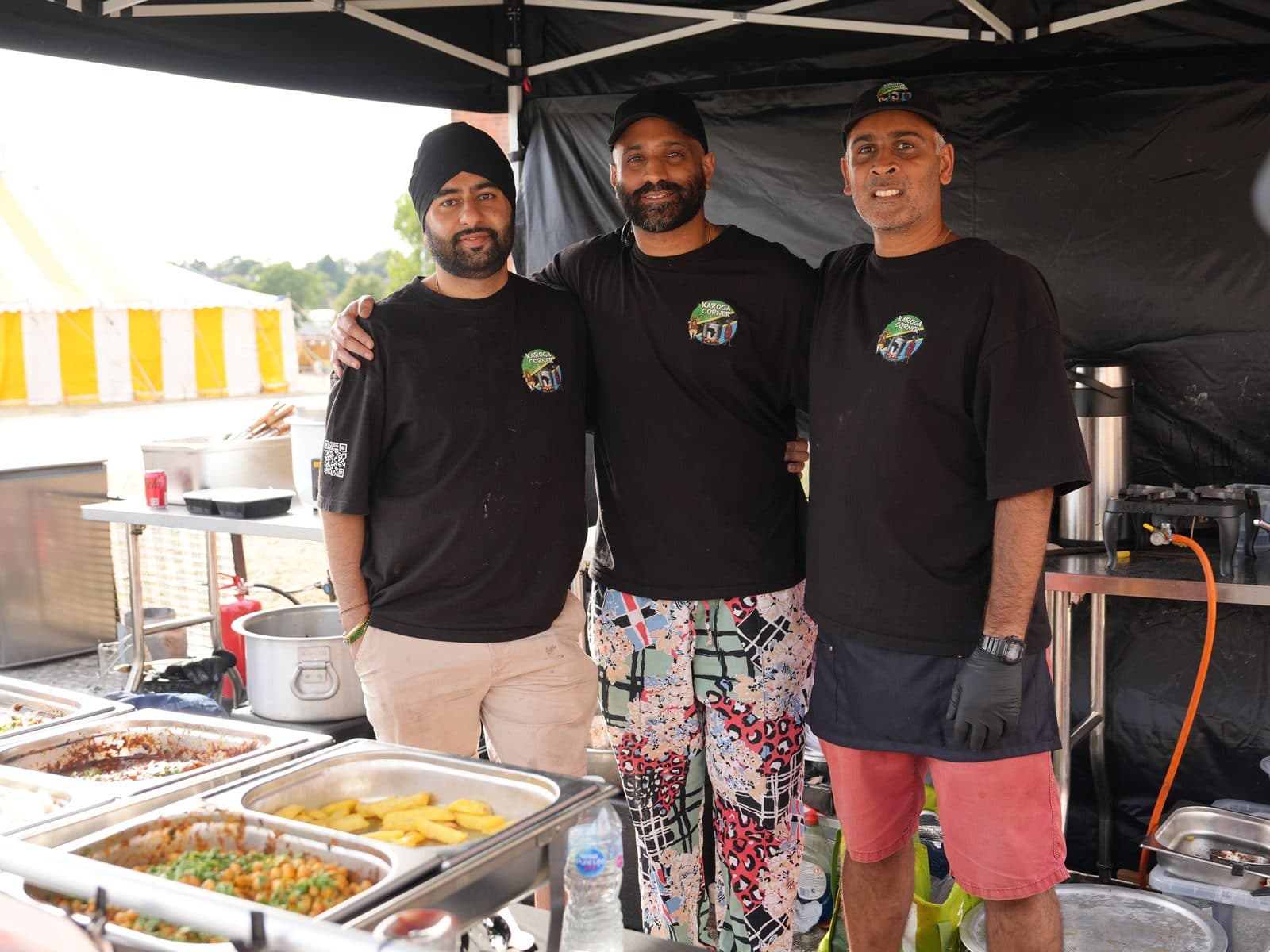 Three of the Karoga Corner team, arms round each other in branded black shirts, behind a counter of full serving trays under the gazebo, with a yellow and white striped marquee and open field behind them.