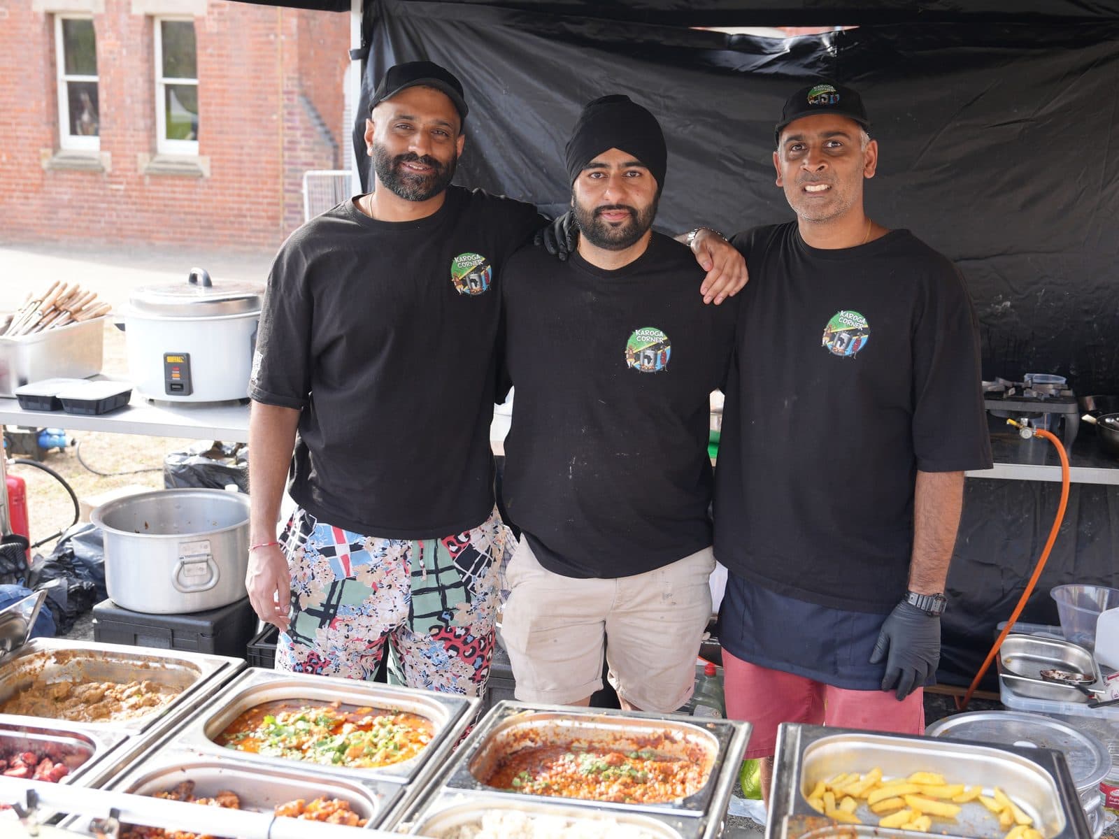 Three of the team shoulder to shoulder behind a full run of serving trays, with the stall's signage and a brick building behind them.