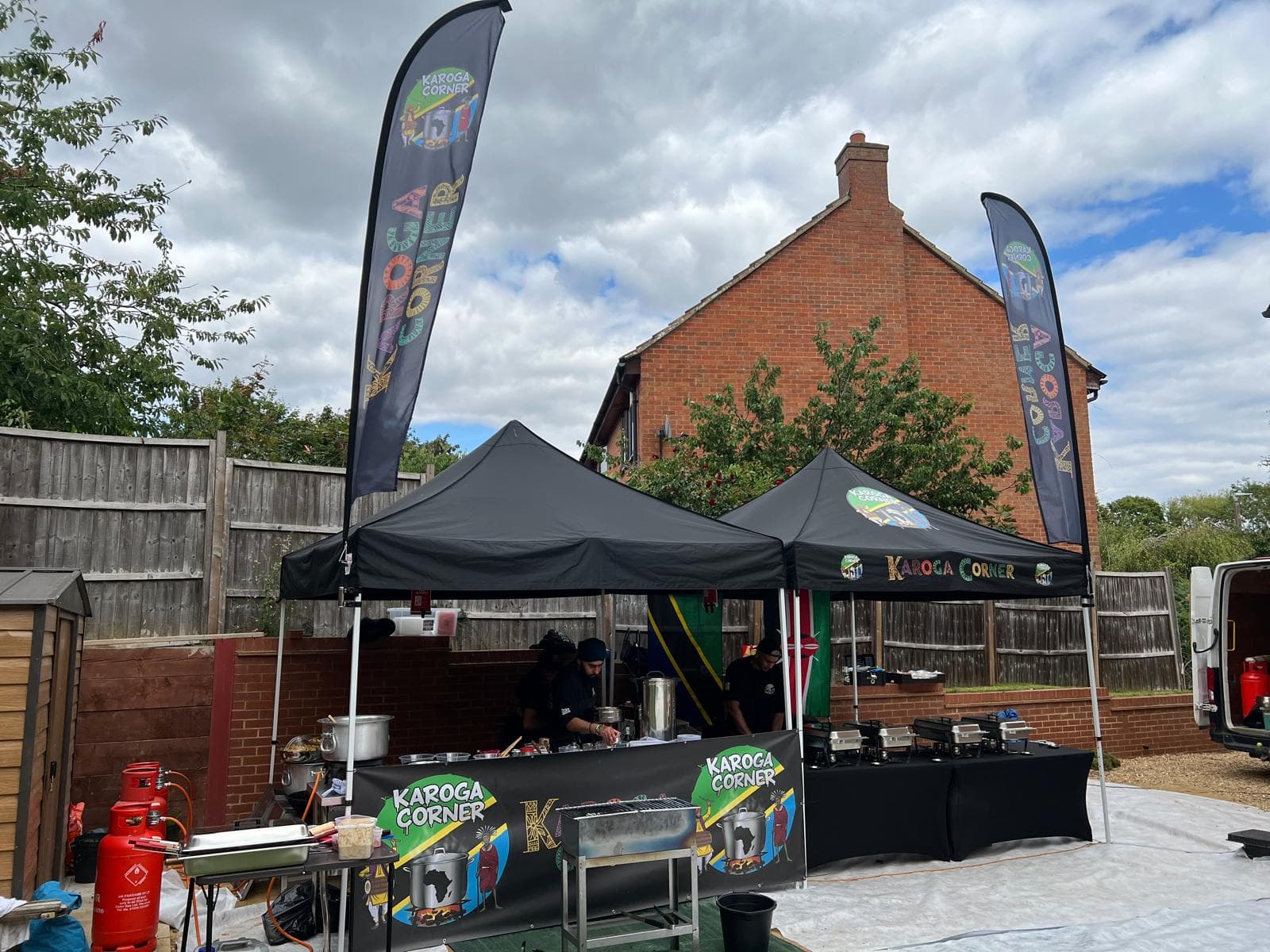 Two black Karoga Corner gazebos and a pair of branded feather flags set up on a driveway for a private booking, with gas burners, a grill and chafing dishes laid out ready.