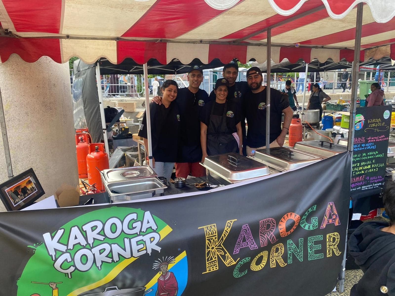 The Karoga Corner team behind their stall at a UK food festival, with the hand-painted Karoga Corner banner across the front.